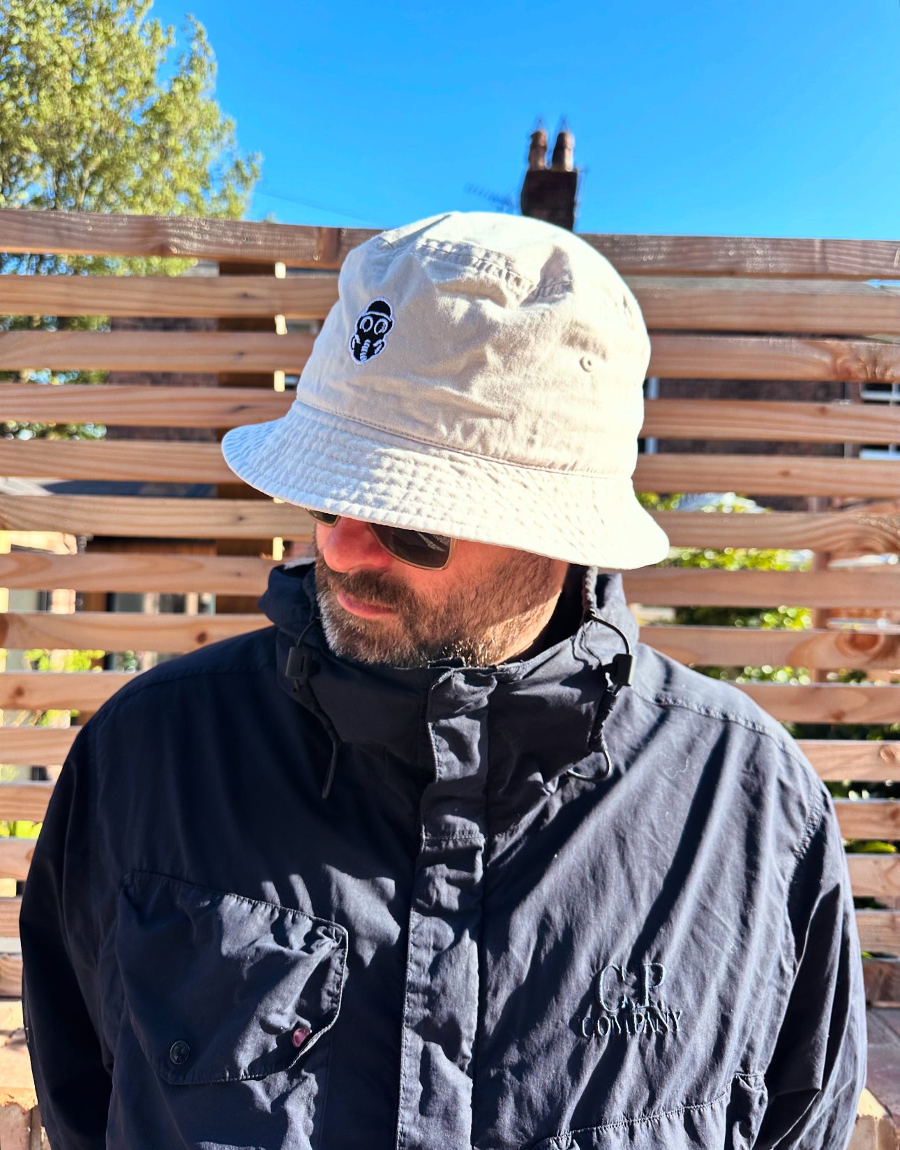 Person wearing a beige bucket hat and dark jacket outdoors with a wooden fence and trees in the background.