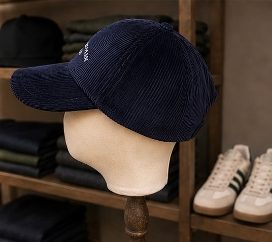 The Saint Talisman Cord Cap - Navy, featuring embroidered detailing and inspired by traditional British hats, is displayed on a beige mannequin head with folded clothing and white sneakers with green stripes in the background.