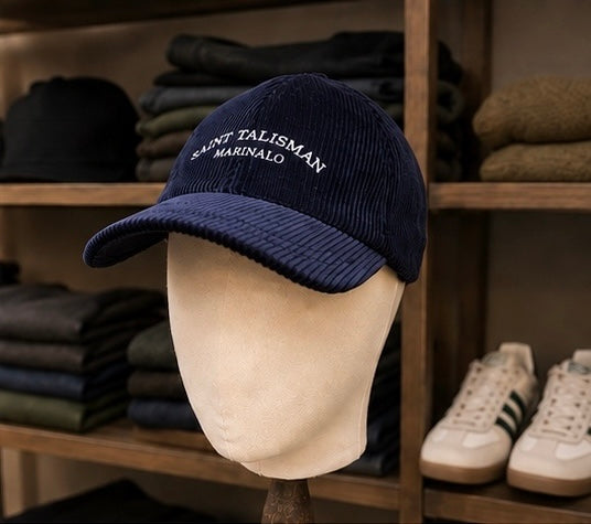 The Saint Talisman Cord Cap - Navy, featuring white embroidered text and crafted from corduroy, is displayed on a mannequin head; folded clothes and white sneakers can be seen arranged on shelves in the background.