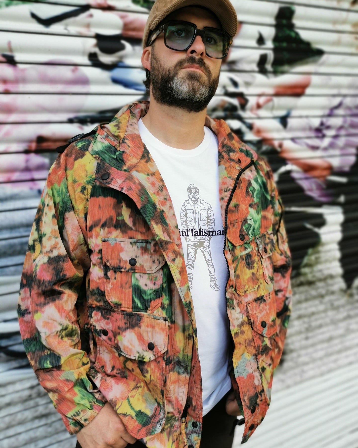 A bearded man in sunglasses and a tan cap stands confidently by a graffiti wall, wearing a bright floral jacket and the Urban Gasman T-Shirt - White, an organic cotton tee with a sketch of a man and the word "Talisman.