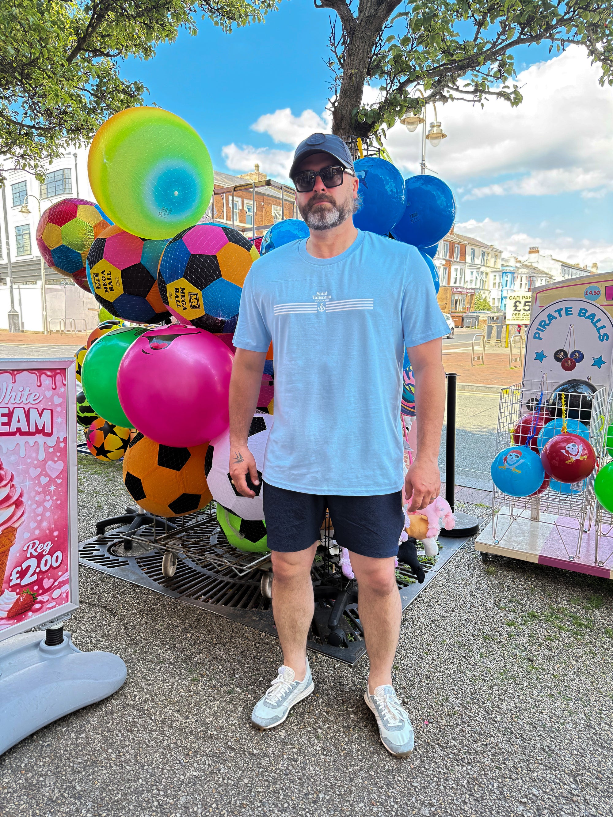 A man wearing a Quartermaster T-Shirt Powder Blue, dark shorts, and a cap stands near a vibrant outdoor market display of large balls. Nearby are signs, a gumball machine, a tree, and buildings in the background.