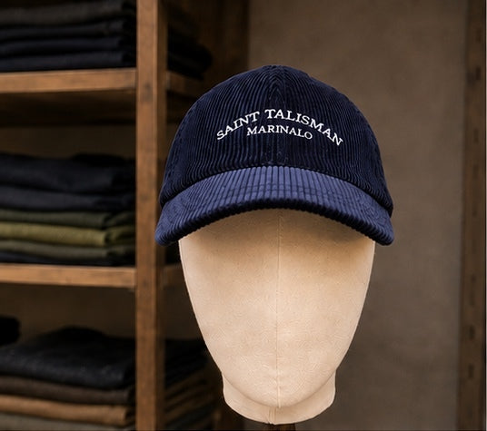 A navy Saint Talisman Cord Cap is showcased on a beige mannequin head, set against shelves lined with folded clothes. This cap features classic white embroidery and evokes the timeless charm of a traditional British hat through its elegant design.