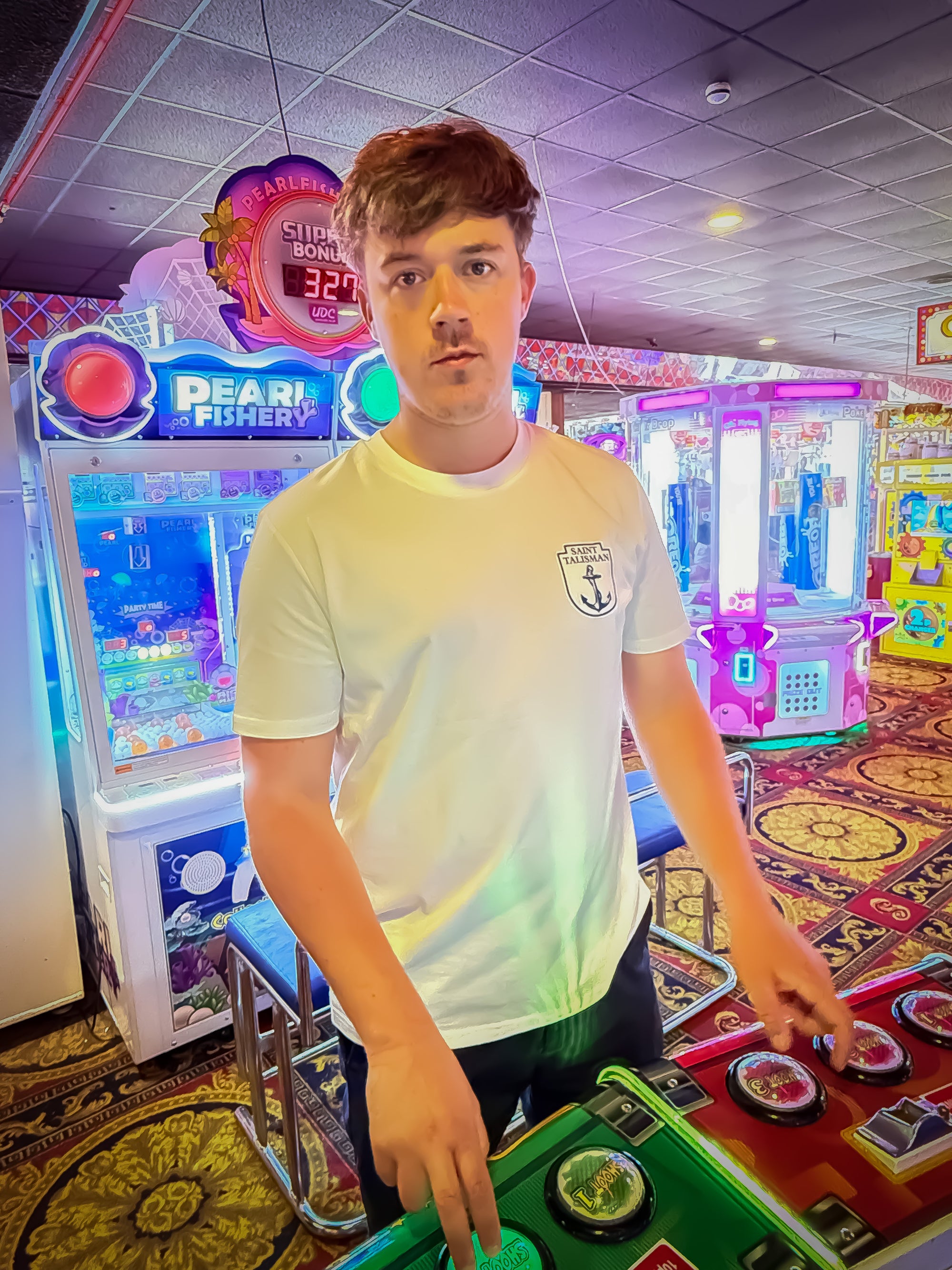 A young man with light brown hair wears THE SHIELD TEE — ANCHOR EDITION in Vintage White as he stands at an arcade game, surrounded by vibrant lights and lively machines.