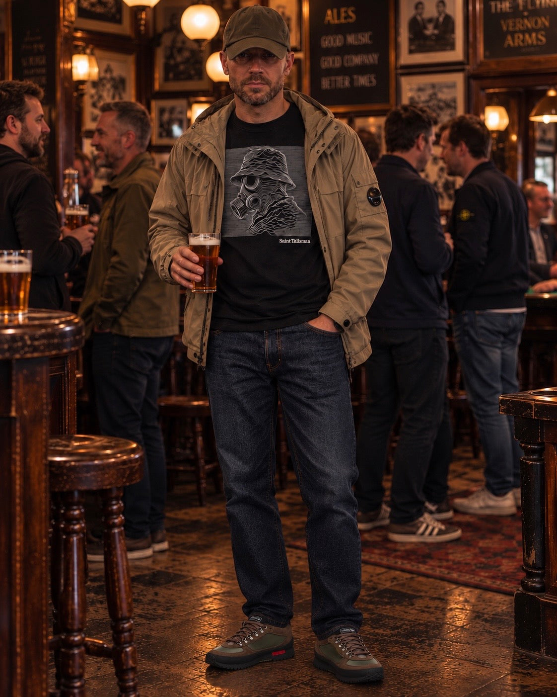 A man wearing a black Unknown Gasman T-shirt - White Design stands in a pub holding a glass of beer.