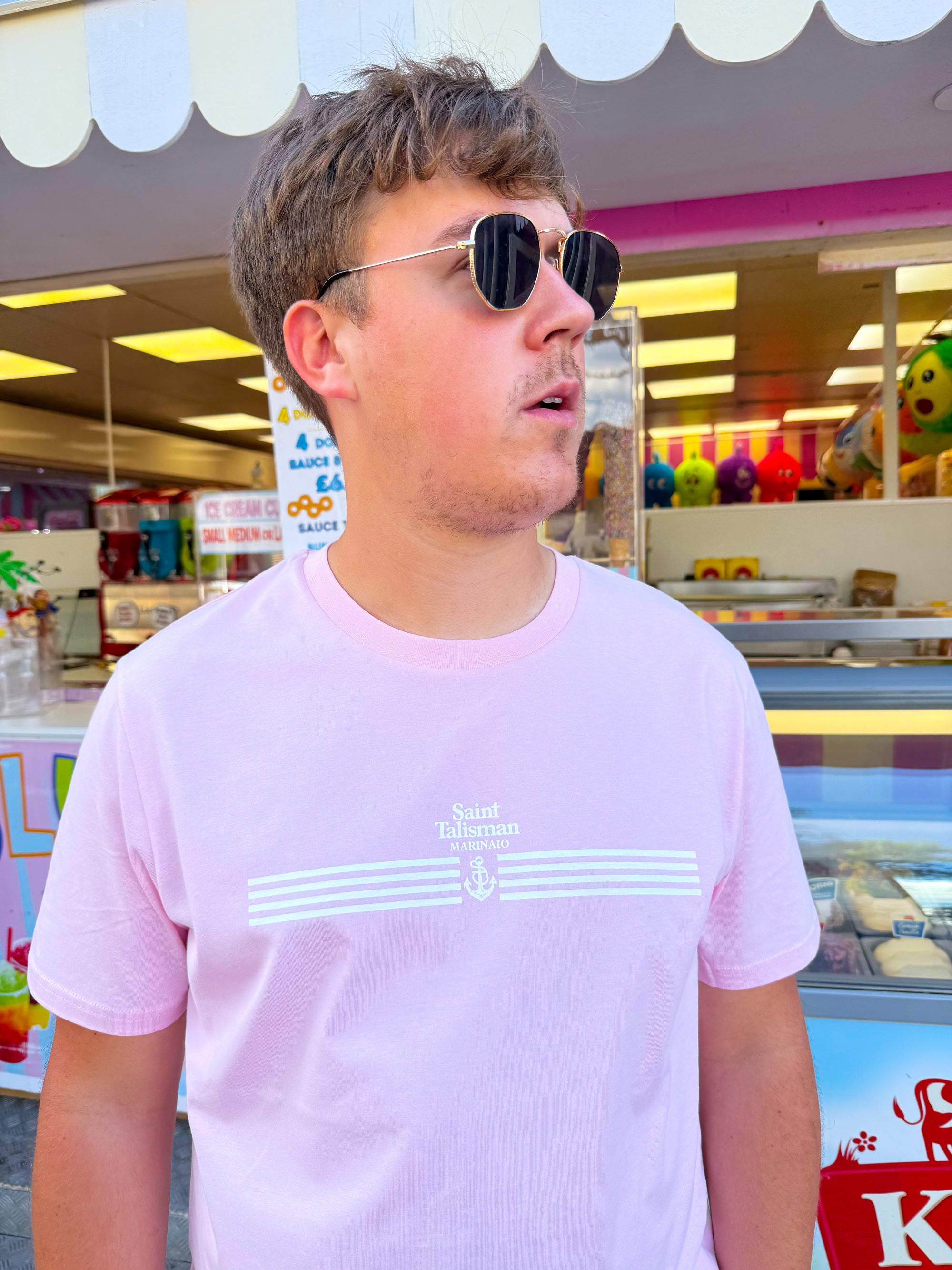 A young man in round sunglasses and a light pink Quartermaster T-Shirt Rose made from organic cotton stands before a colorful food stall, glancing to the side with his mouth slightly open.