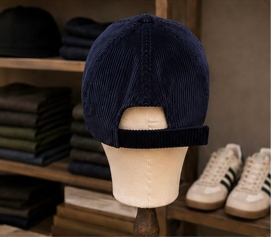 A Saint Talisman Cord Cap in navy is displayed on a beige mannequin head, with shelves of folded clothes and a pair of white sneakers visible in the background.
