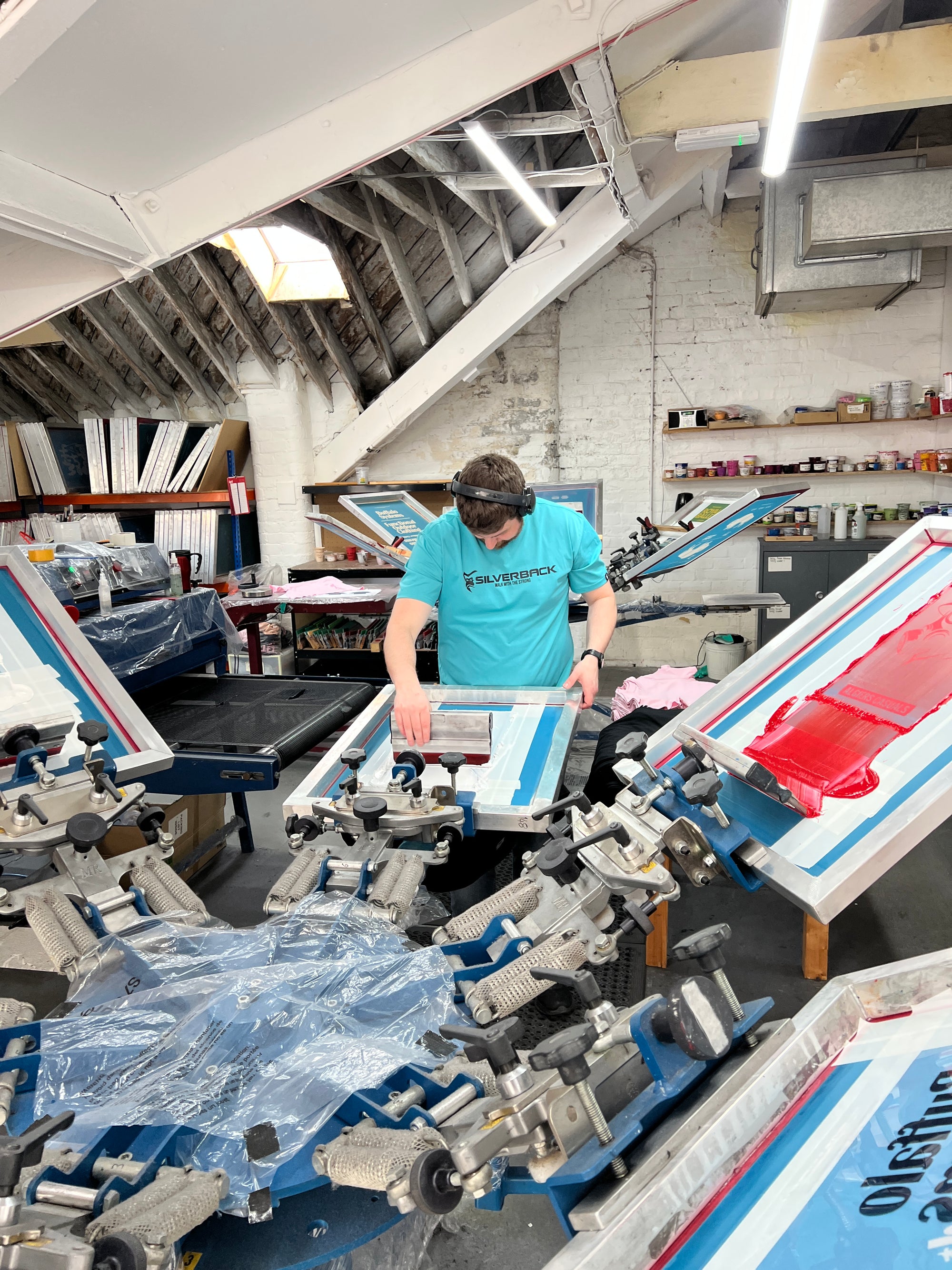 A person in safety goggles and a blue SAINT TALISMAN x ALGIERS CASUALS t-shirt screen prints in a bright workshop, surrounded by presses and supplies—a relaxed, creative scene inspired by football culture.