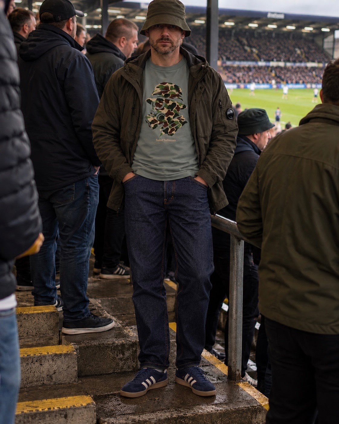 A person wearing a grey CAMO GAS MASK T-SHIRT with a camouflage gas mask graphic, an olive green jacket, and a bucket hat while standing in a stadium.