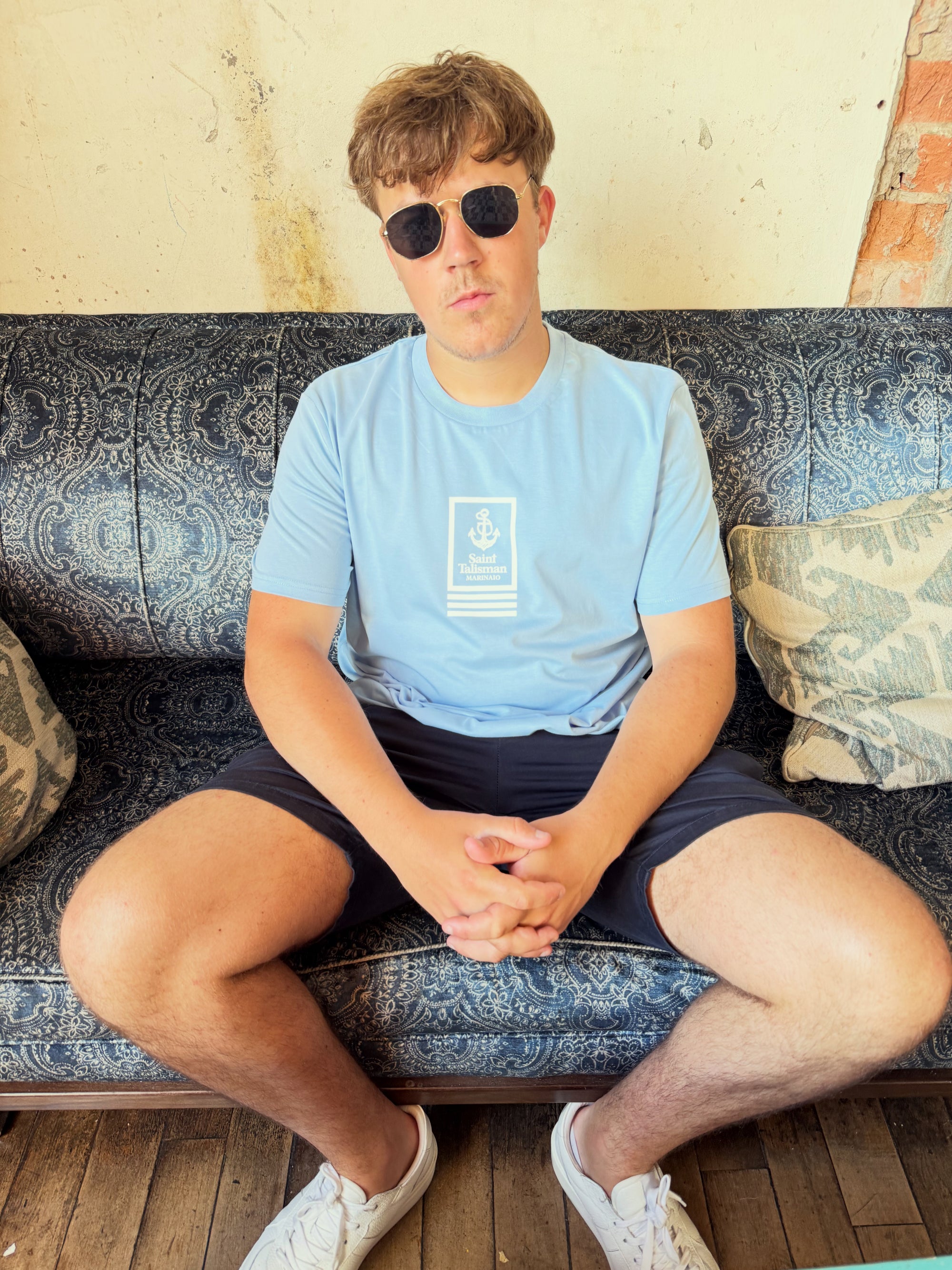 A young man wearing the Union T-Shirt in powder blue and sunglasses sits on a patterned sofa with decorative cushions, hands clasped, looking at the camera against a textured wall.