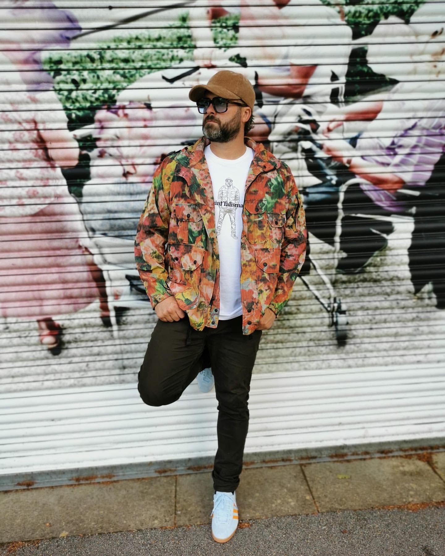 A man wears the Urban Gasman T-Shirt in white, paired with a medium-fit floral jacket, black pants, white sneakers, brown cap, and sunglasses as he poses casually against a mural-covered corrugated metal wall with one leg bent.