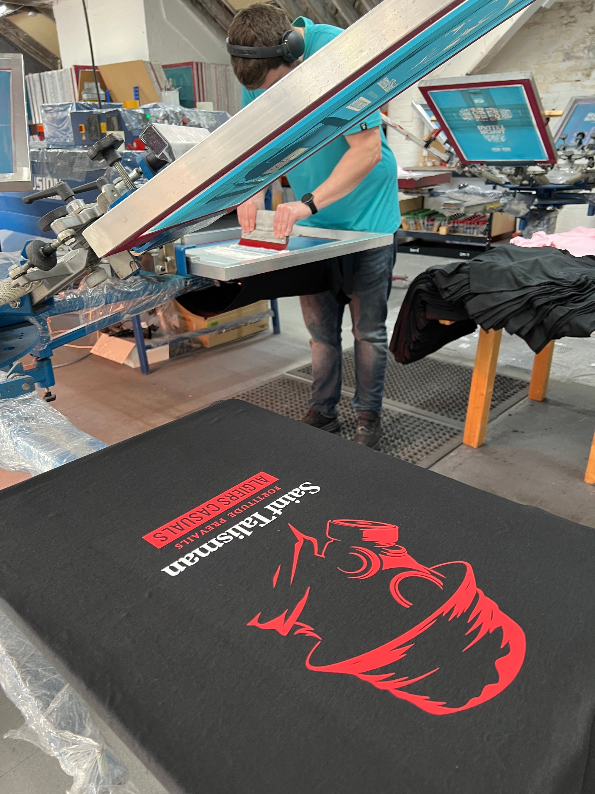 Amid equipment and piles of clothing, someone uses a screen printing machine to create a black SAINT TALISMAN x ALGIERS CASUALS T-shirt, boldly blending football culture with fashion in vivid red and white.
