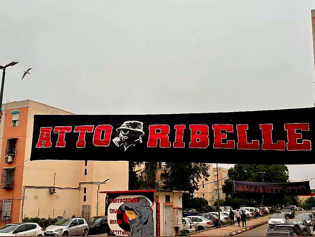 A black banner with bold red and white text reading "SAINT TALISMAN x ALGIERS CASUALS" hangs between apartment buildings. A sunglasses-wearing face appears amid the words, referencing football culture. Cars and a bird in the sky are visible below.