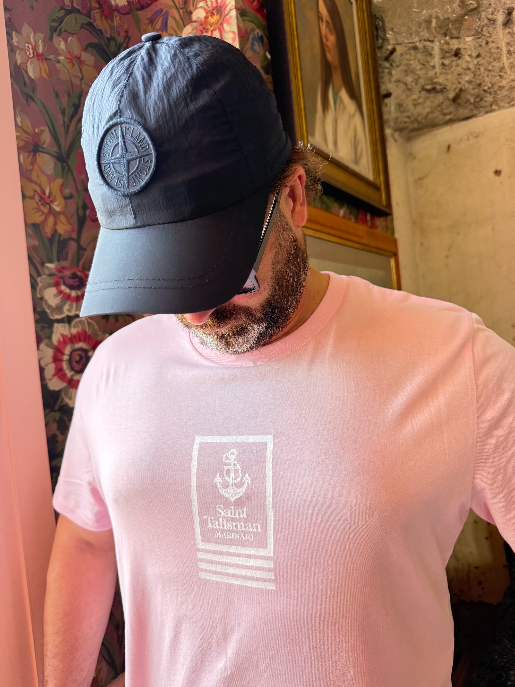 A bearded man in sunglasses, a black cap, and the Union T-Shirt Rose looks down while standing in a room with floral wallpaper and framed artwork behind him.