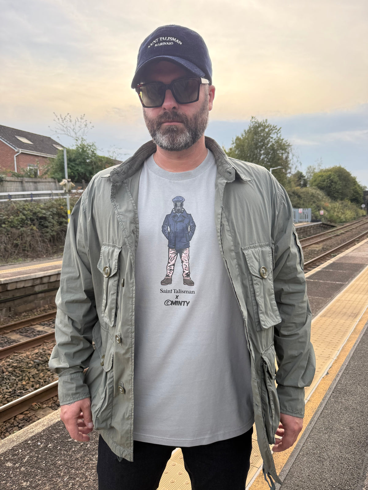 A man in sunglasses, a dark cap, and a green jacket stands on a train platform, wearing the ST x Minty Marinaio Gasman T-Shirt featuring a parka-clad figure with the text Saint Talisman.
