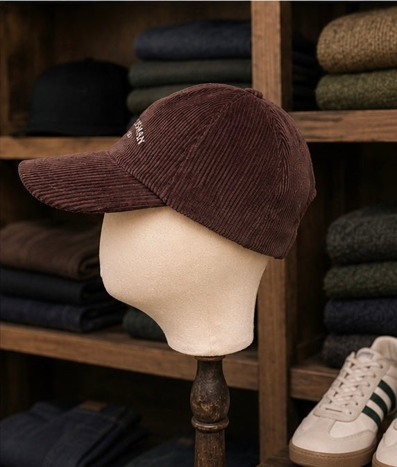 The Saint Talisman Cord Cap - Brown is stylishly perched atop a mannequin head in a clothing store display, with folded garments and white sneakers featuring green stripes visible in the background.