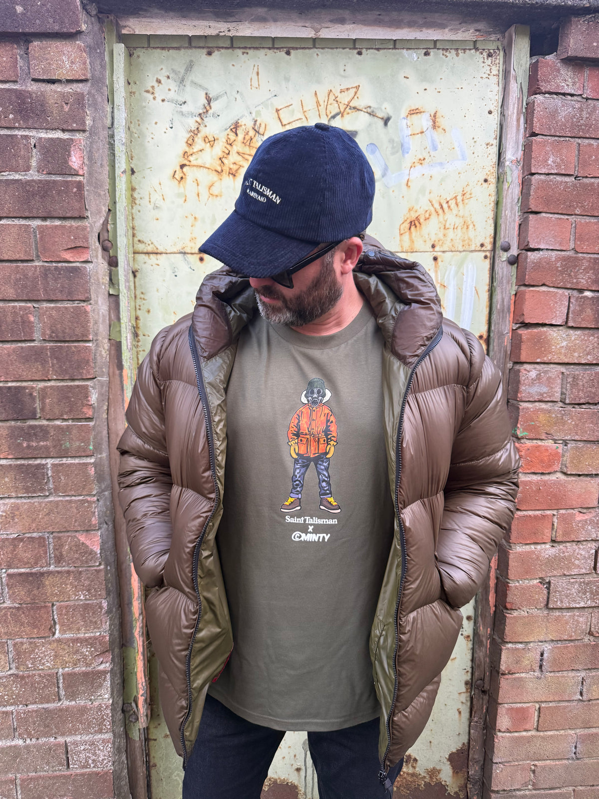 A man in a brown puffer jacket, ST x Minty Alpine Gasman T-Shirt, and black cap stands before a graffiti-covered door, looking down with his hands in his jacket pockets.