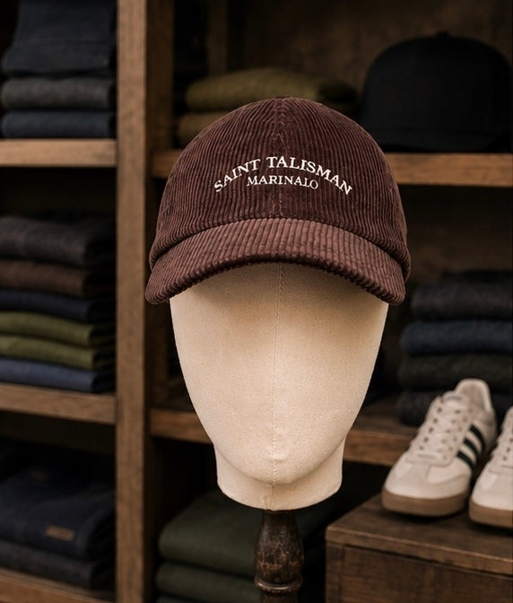 A Saint Talisman Cord Cap - Brown is displayed on a mannequin head, featuring “MARINAIO” embroidered on the front and surrounded by folded clothes and a sneaker in the background, blending modern style with the classic charm of a traditional British hat.