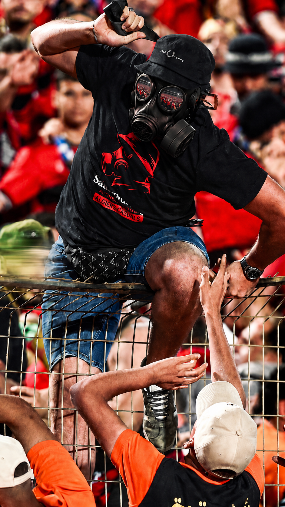 Wearing a SAINT TALISMAN x ALGIERS CASUALS black shirt and gas mask, a person climbs over a metal fence, reaching for help amid a crowd where football culture is visible in the background.