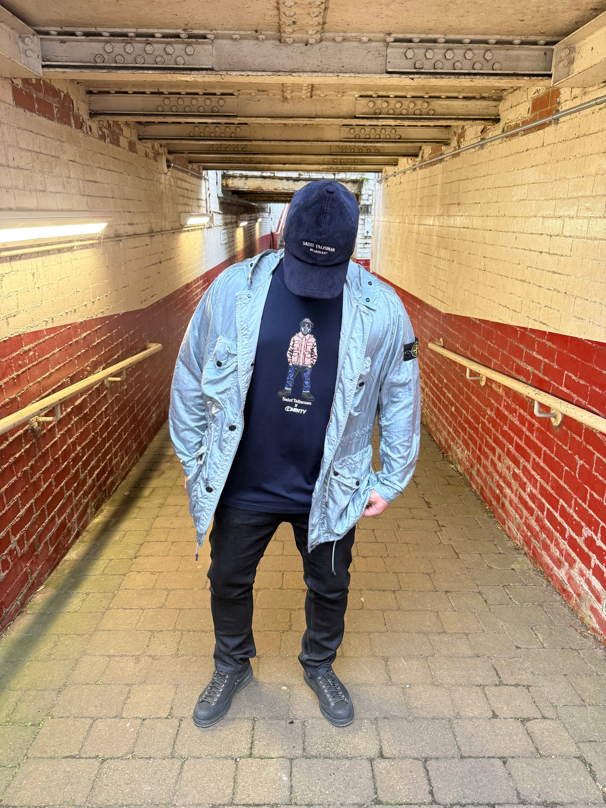 Wearing the ST x Minty Urban Gasman T-Shirt, a person in a blue jacket, black pants, and black cap stands in a red-and-white brick tunnel with hands in pockets, radiating cool confidence.