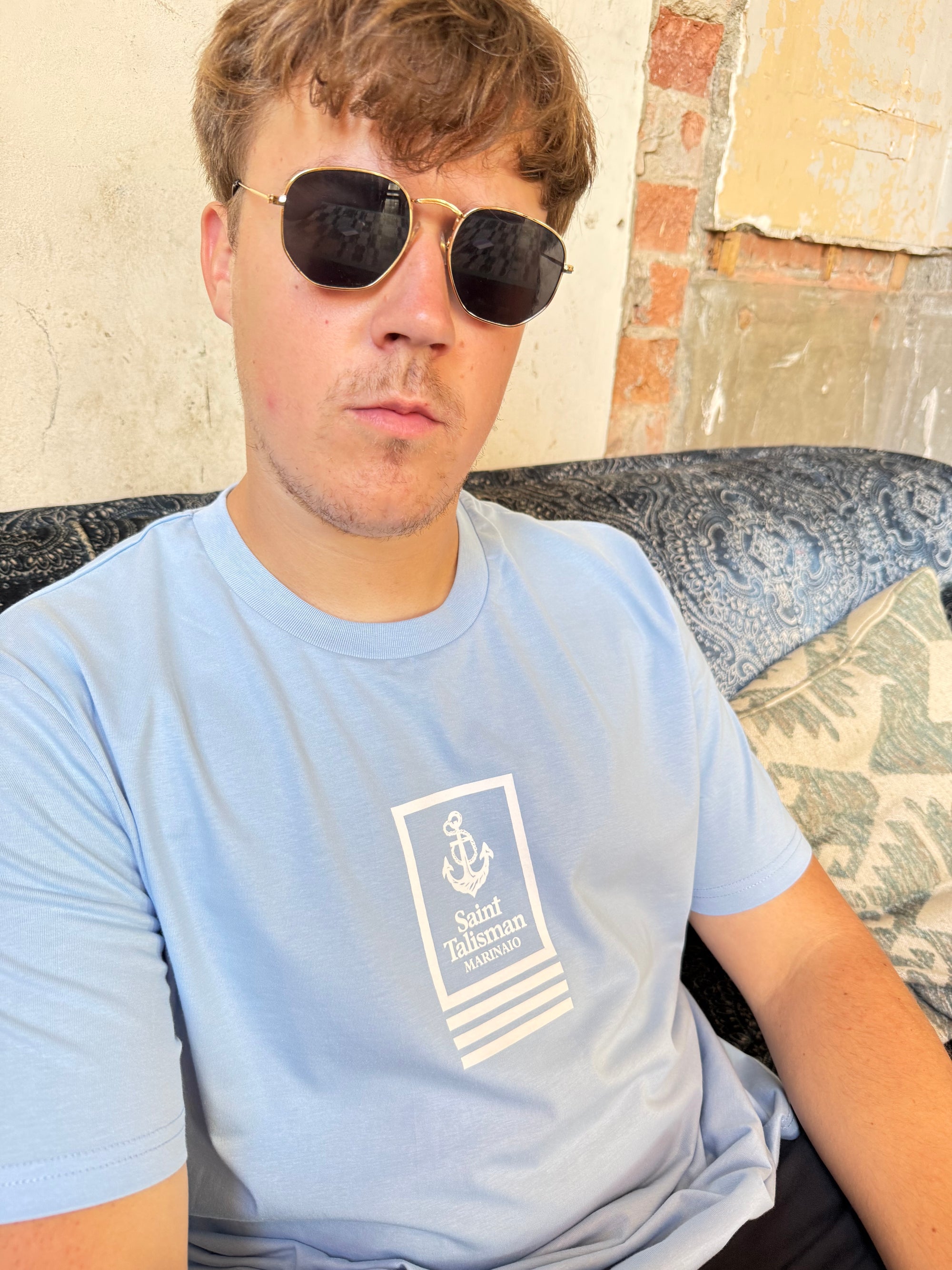 A young man in sunglasses and a powder blue Union T-Shirt sits on a patterned couch against a rustic wall with peeling paint and exposed bricks, creating a relaxed vibe.