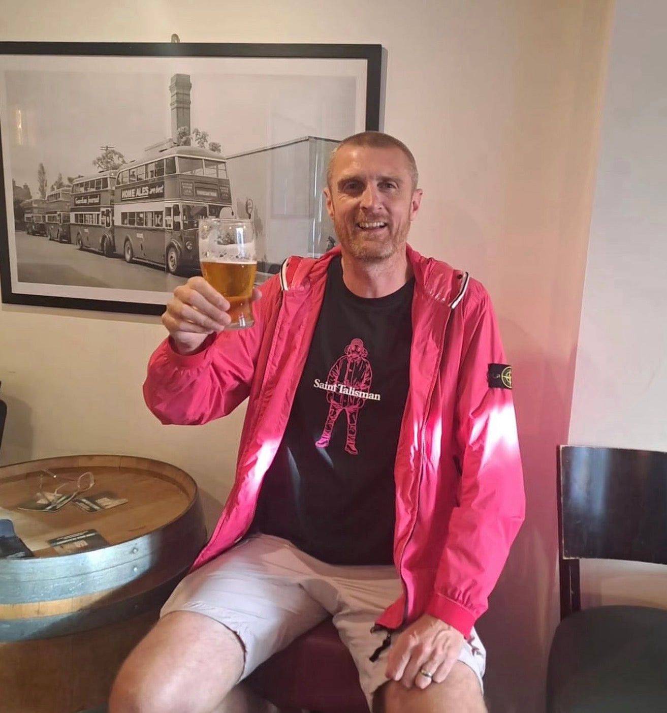 A man wearing the Alpine Gasman T-Shirt - Black & Pink sits indoors, smiling and holding a pint of beer. Behind him is a framed black-and-white bus photo, and a round wooden table is beside him.