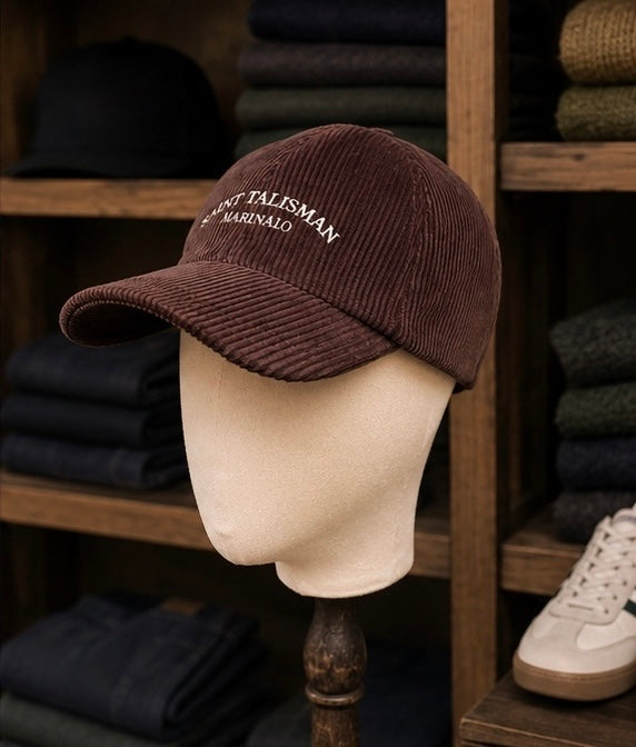 The Saint Talisman Cord Cap - Brown, featuring white embroidered text, is displayed on a beige mannequin head. In the background, wooden shelves hold folded clothes and a pair of white sneakers.
