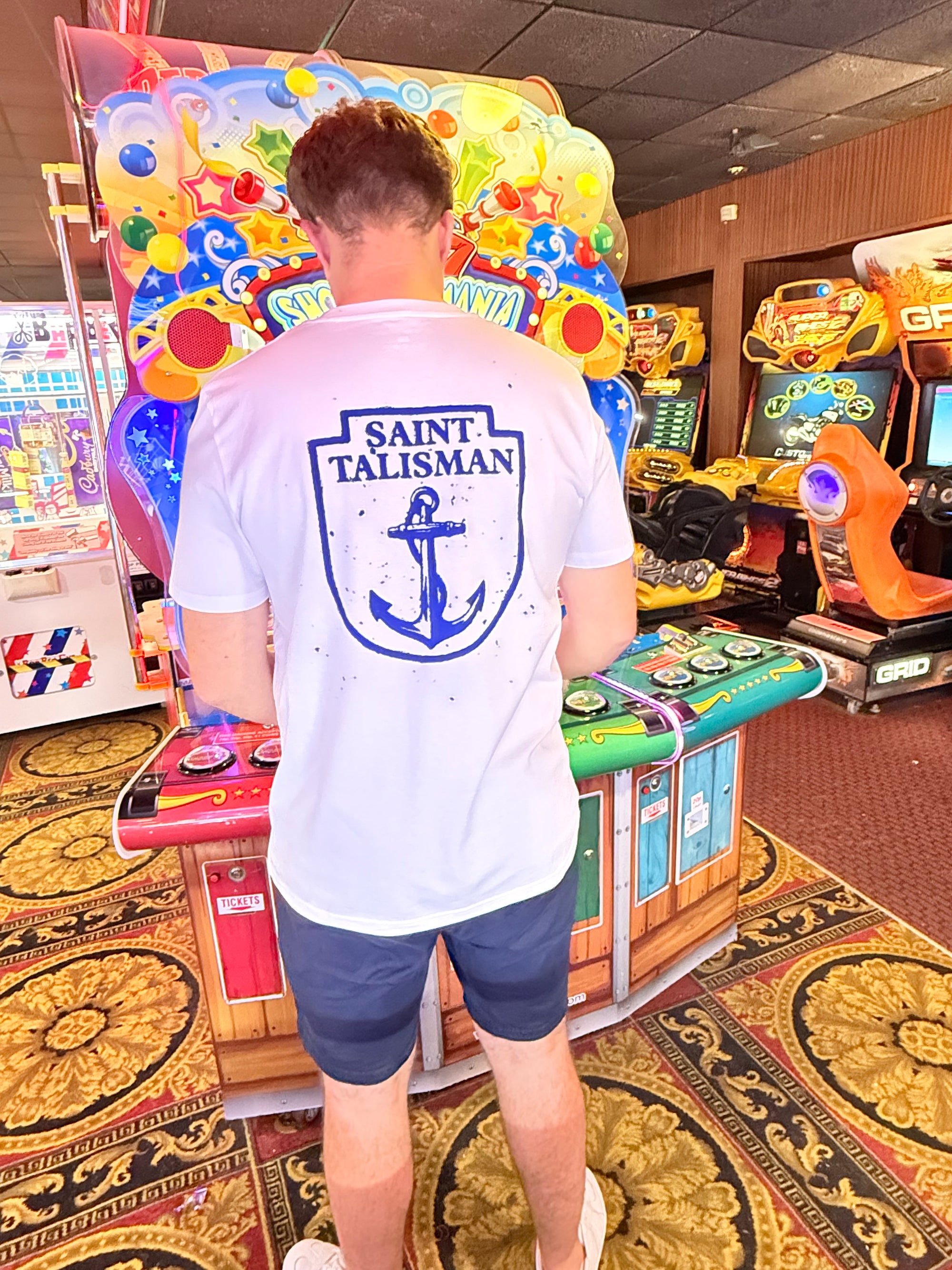 A person with light brown hair, wearing a vintage white THE SHIELD TEE — ANCHOR EDITION and blue shorts, stands at a colorful arcade game in a brightly lit arcade room.