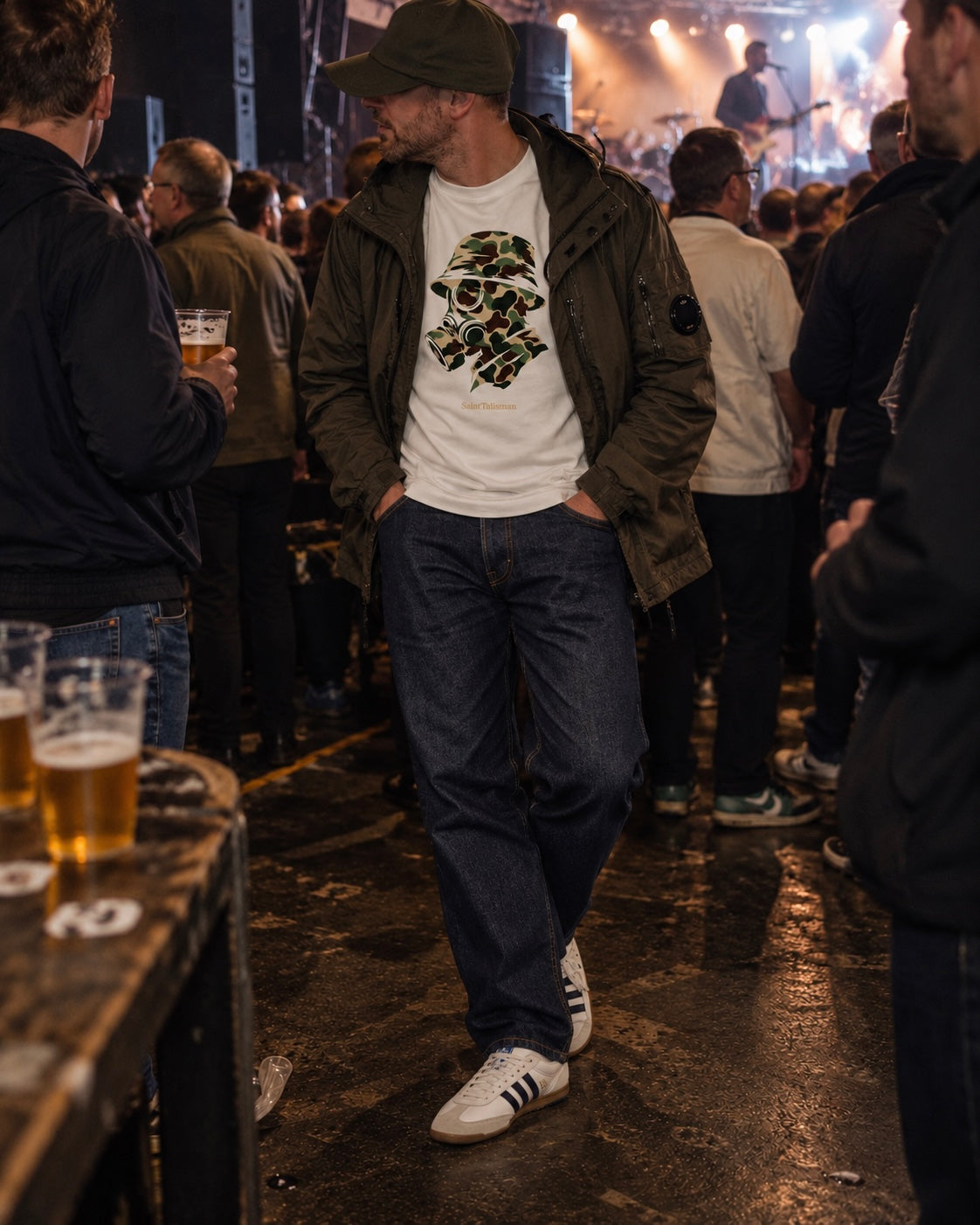 A man in a white CAMO GAS MASK T-SHIRT layered under an olive green jacket, standing in a crowded venue.