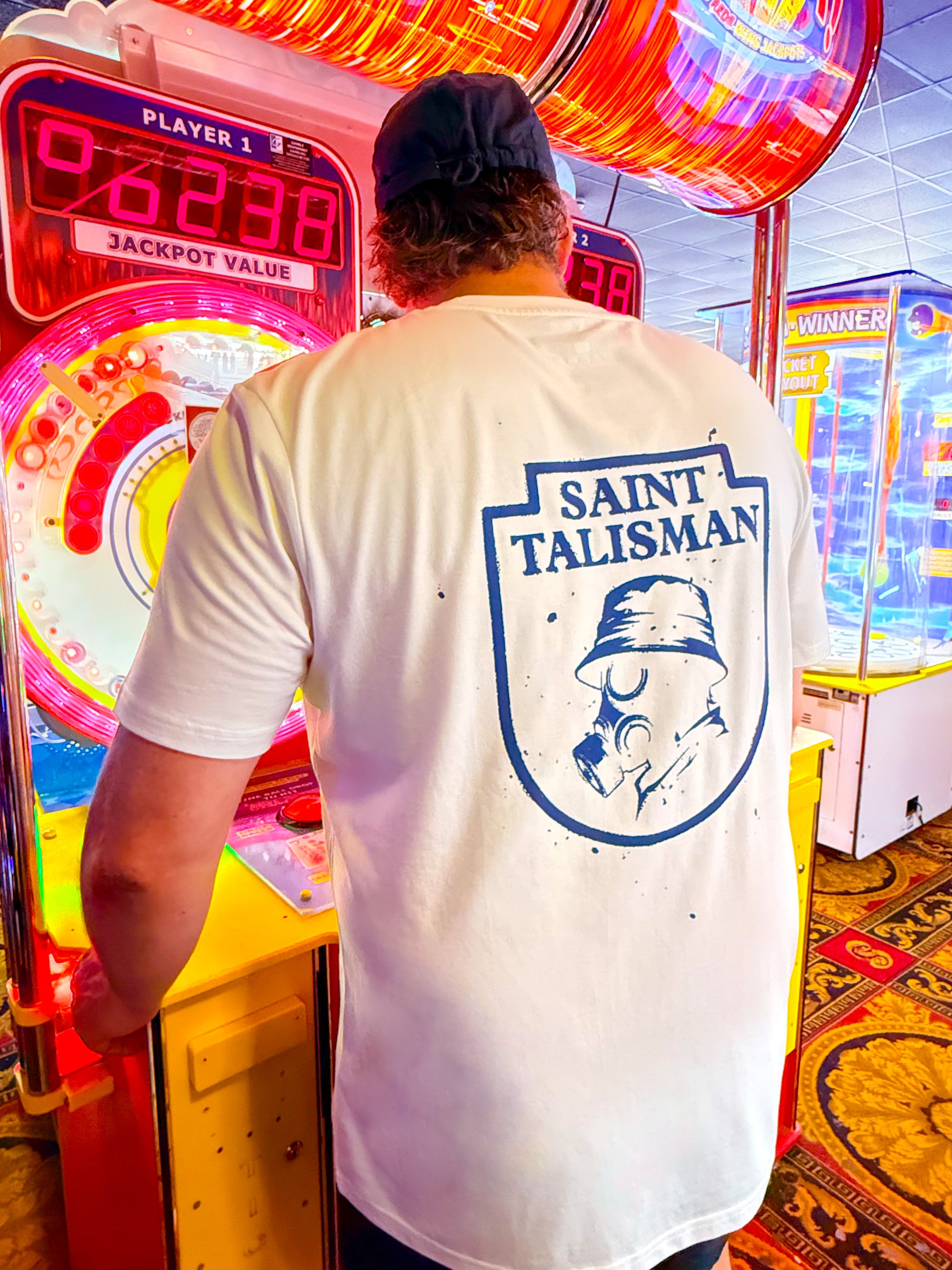 A person in a Vintage White THE SHIELD TEE — GAS MASK EDITION stands at an arcade game machine, surrounded by bright lights and colorful designs.