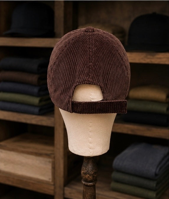 The Saint Talisman Cord Cap - Brown is displayed from the back on a beige mannequin head, with folded clothes neatly stacked on wooden shelves in the background.