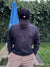 Wearing a black cap and the MK2 Danforth Embroidered Marinaio Sweatshirt in Stealth, featuring an anchor design, a person stands outdoors by a wooden picnic table, embodying classic British casual style amid greenery and a blue umbrella in the background.