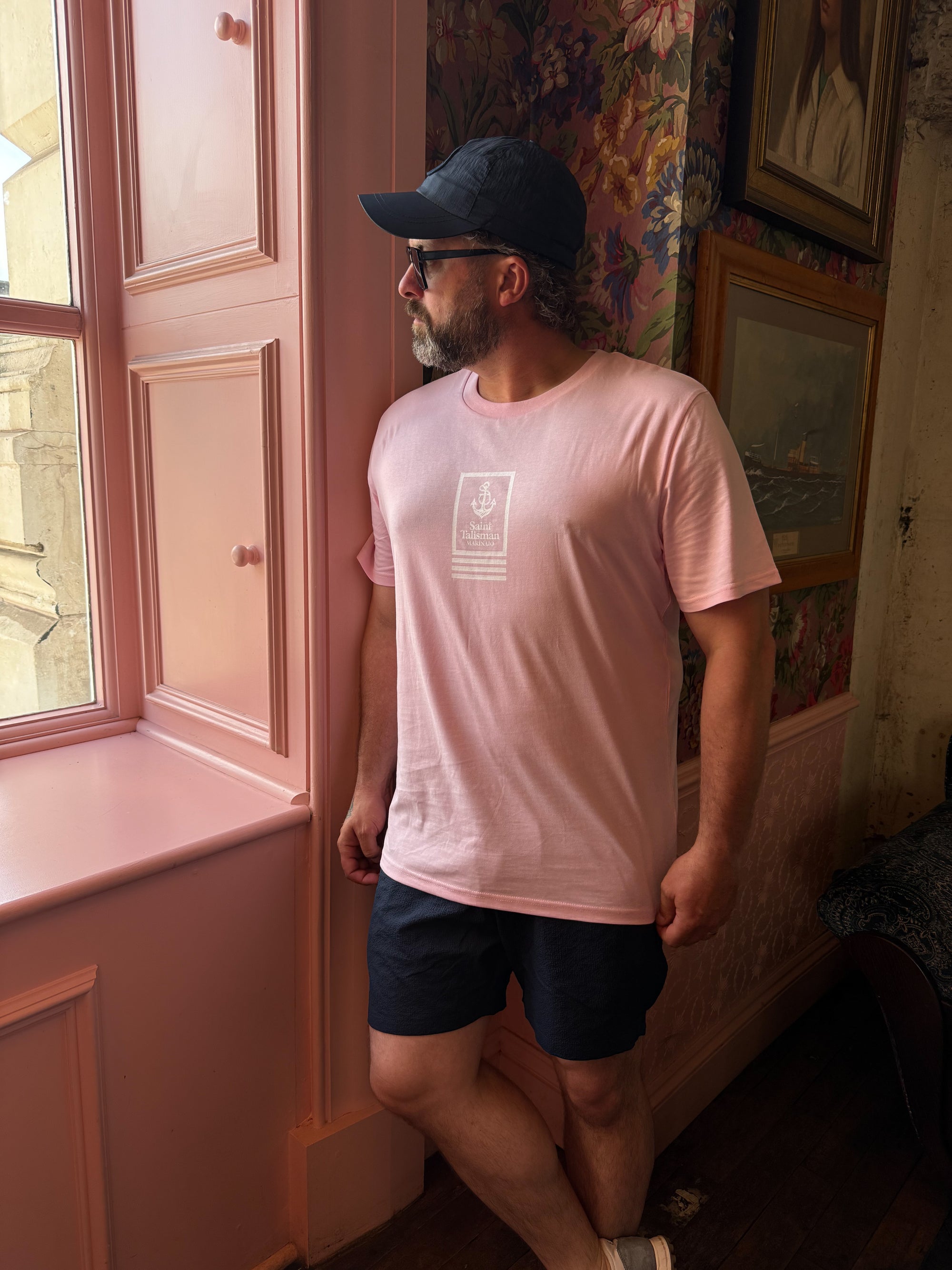 A man wearing a Union T-Shirt Rose and dark shorts stands by a pink window, looking outside. He has a beard, sports sunglasses and a black cap, in a floral wallpapered room with framed pictures.
