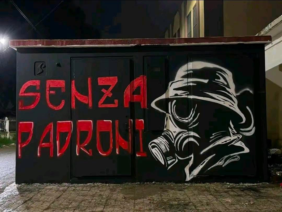 The SAINT TALISMAN x ALGIERS CASUALS mural features "Senza Padroni" in red beside a white outline of a bucket-hatted, gas-masked figure on a black wall, evoking football culture in an outdoor nighttime setting.