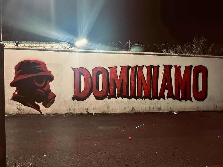 A nighttime mural for SAINT TALISMAN x ALGIERS CASUALS shows “DOMINIAMO” in bold red on a white wall, with a figure in a red hat and gas mask beside barbed wire—capturing the intensity of football culture.