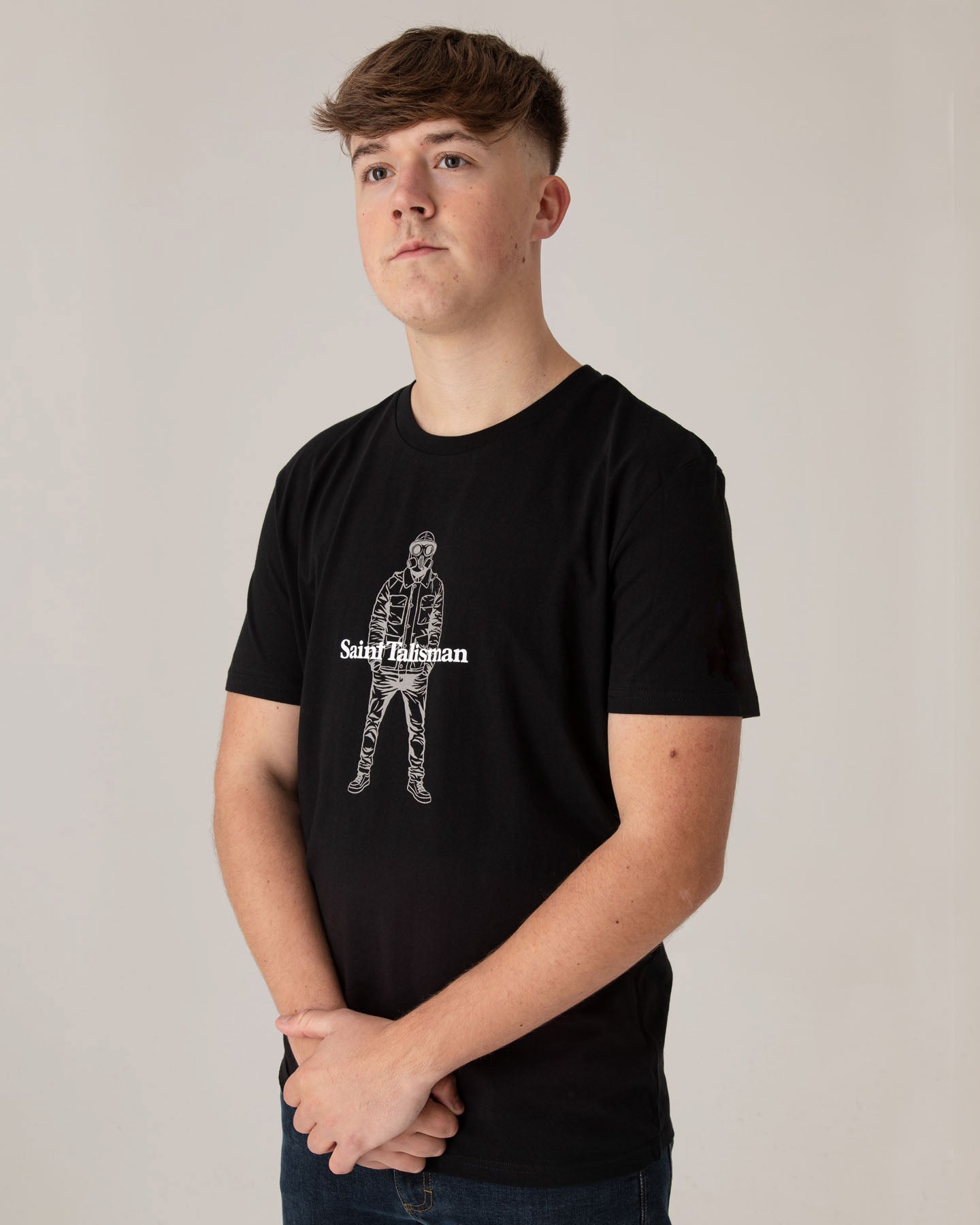 Young man with short brown hair wears a medium fit black Urban Gasman T-Shirt—crafted from organic cotton, featuring a white line drawing of a person—standing against a plain light background with hands clasped in front.