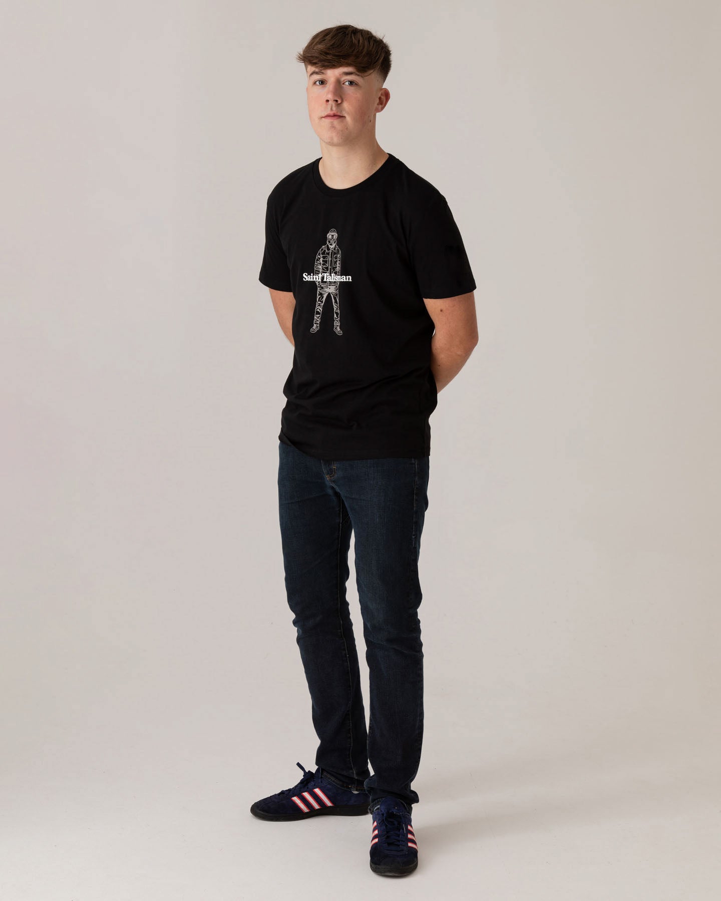 A young man with short brown hair stands against a plain light background, wearing the Urban Gasman T-Shirt - Black & Grey, dark jeans, and black sneakers with red stripes. He has his hands behind his back and is looking at the camera.