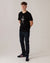 A young man with short brown hair stands against a plain light background, wearing the Urban Gasman T-Shirt - Black & Grey, dark jeans, and black sneakers with red stripes. He has his hands behind his back and is looking at the camera.