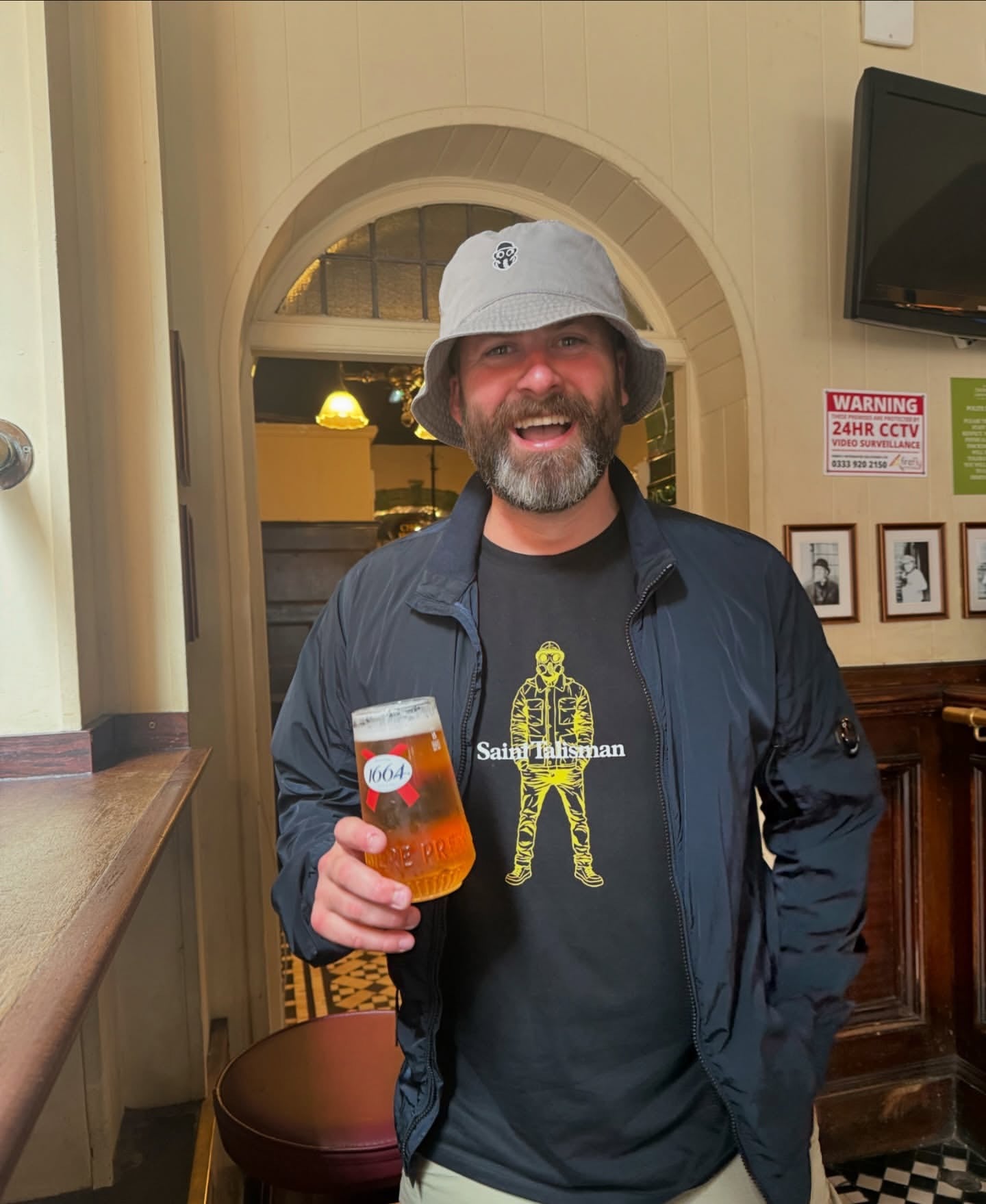 A smiling bearded man in a gray bucket hat and black jacket sports the Urban Gasman T-Shirt - Black & Yellow while holding a pint of beer inside a pub, with signs and a TV visible on the wall behind him.