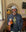 A smiling bearded man in a gray bucket hat and black jacket sports the Urban Gasman T-Shirt - Black & Yellow while holding a pint of beer inside a pub, with signs and a TV visible on the wall behind him.