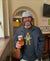 A smiling bearded man in a gray bucket hat and black jacket sports the Urban Gasman T-Shirt - Black & Yellow while holding a pint of beer inside a pub, with signs and a TV visible on the wall behind him.