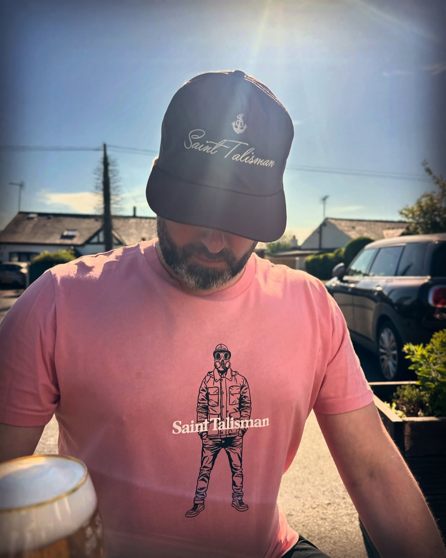 A man sits outdoors in a medium fit, organic cotton Urban Gasman T-Shirt - Pink with a graphic of a person in a jacket and cap, paired with a dark Saint Talisman cap. A drink is on the table, with cars and houses in the background.