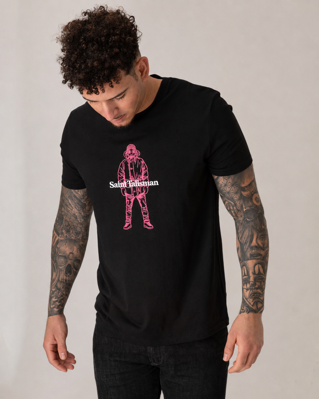 A man with curly hair and tattooed arms looks down while wearing a medium fit Alpine Gasman T-Shirt in black, made from organic cotton, featuring a pink graphic and "Saint Talisman" text on the front.