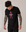 A man with curly hair and tattooed arms looks down while wearing a medium fit Alpine Gasman T-Shirt in black, made from organic cotton, featuring a pink graphic and "Saint Talisman" text on the front.