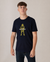 A young man with short brown hair stands against a plain background, wearing the Alpine Gasman T-Shirt in navy & yellow, featuring a graphic and text on the front. He looks slightly to the side.