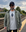 A man in sunglasses, a dark cap, a green jacket, and the ST x Minty Marinaio Gasman T-Shirt stands on a train platform with tracks and trees under a cloudy sky.